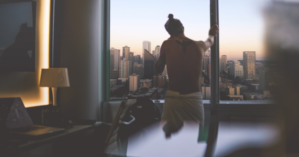 Silhouette of a young man standing at a window looking out at a city skyline at dusk with warm golden light